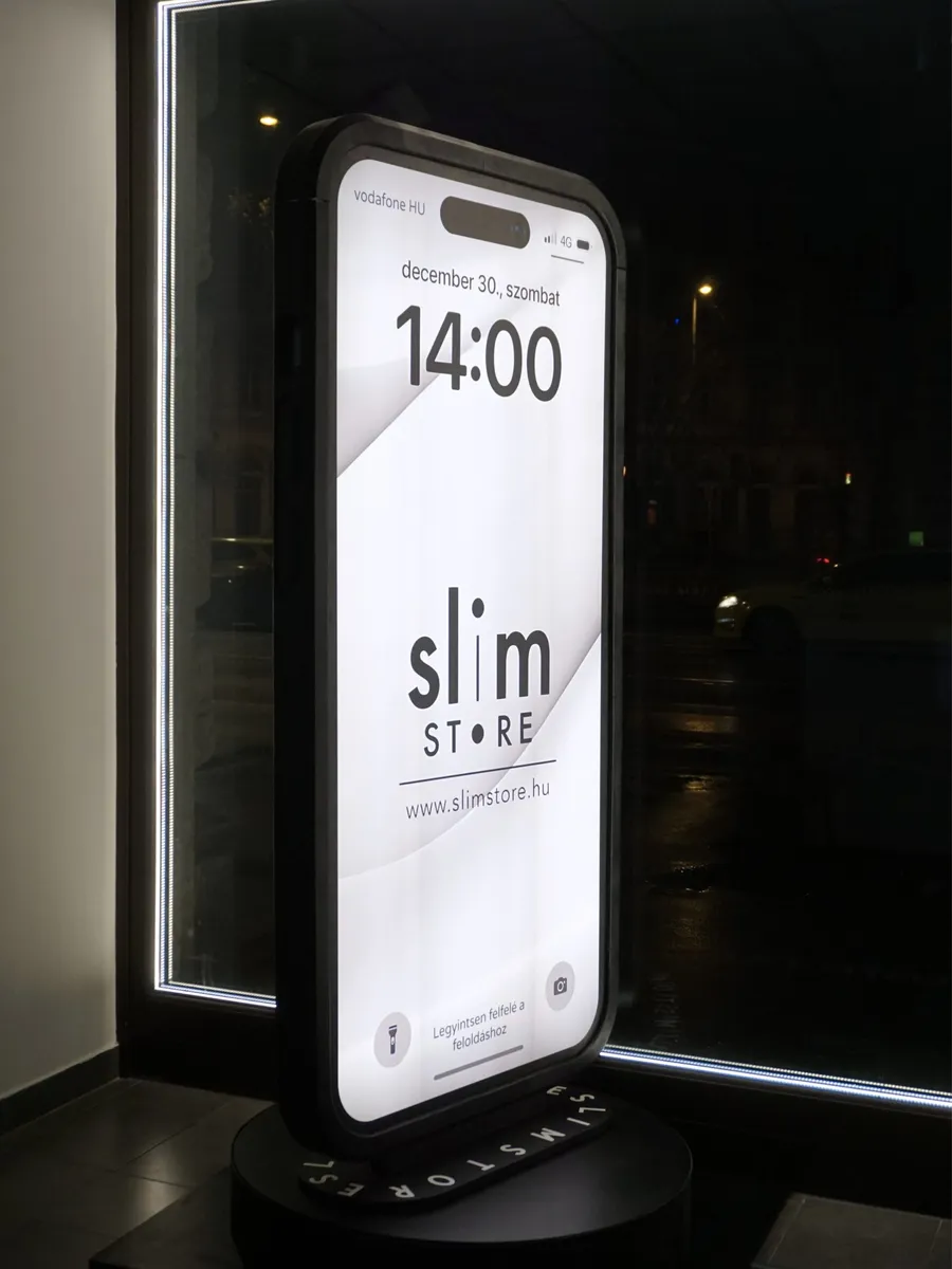 Az óriás iPhone-replika kijelzős oldala este, forgó talpon a Slim Store kirakatában